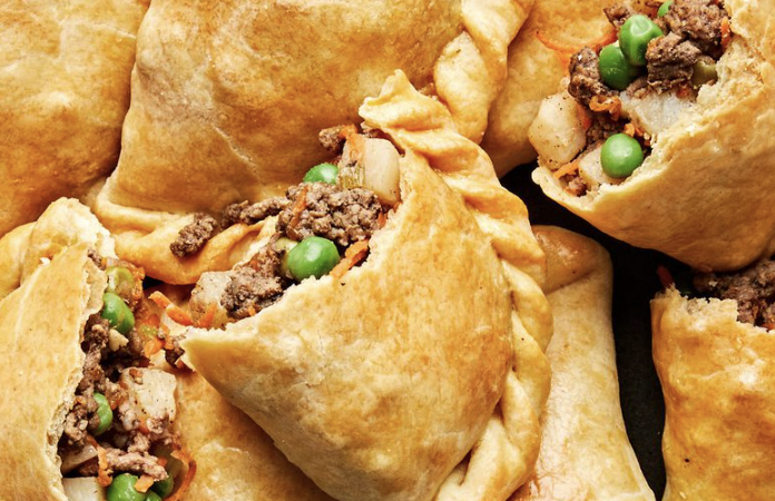 Golden-brown baked pastries filled with ground beef, green peas, potatoes, and carrots, some cut open to reveal the savory filling.