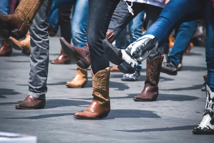 A group of people dressed in jeans and cowboy boots raises their legs while performing a coordinated dance routine on a concrete surface.