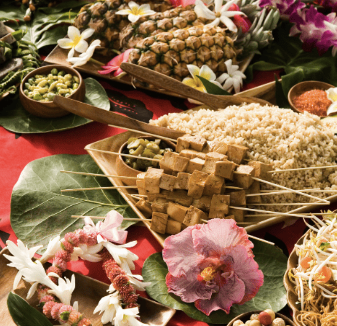 A festive spread of tropical-themed food, including tofu skewers, rice, edamame, pineapple, and garnishes, displayed on large green leaves with flowers and leis for decoration.