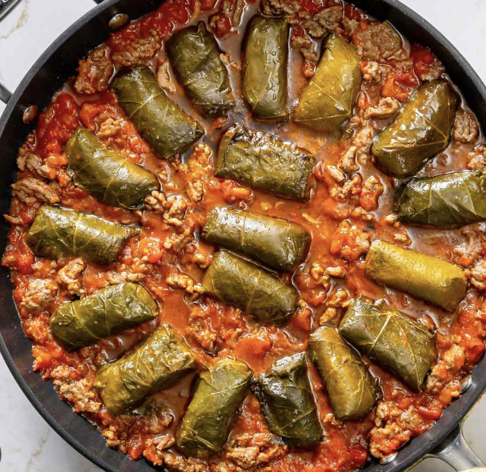 Grape leaves stuffed with rice and meat are arranged in a skillet with a tomato-based sauce and ground meat, all cooked together and garnished with sauce.