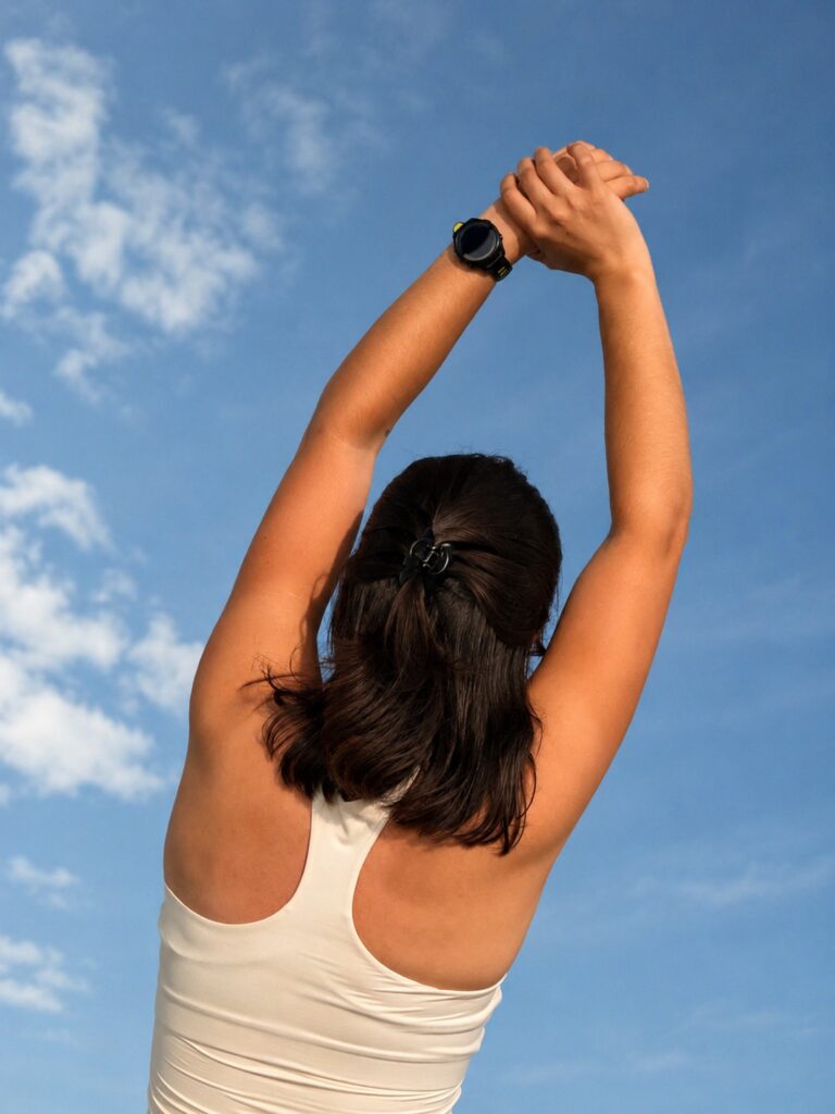 A person with dark hair in a ponytail stretches their arms overhead outdoors under a blue sky with scattered clouds, wearing a light tank top and a watch.