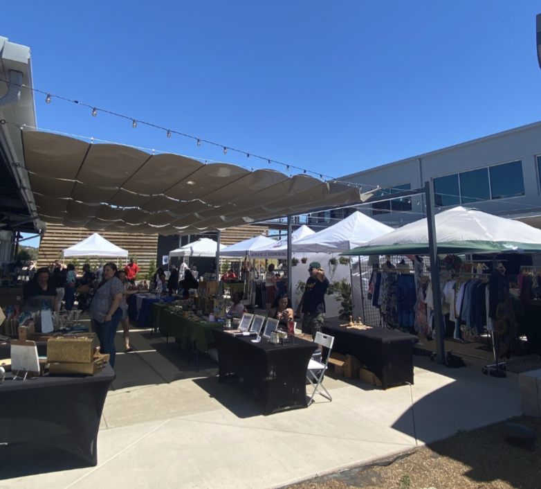 Outdoor market with people browsing tables of various goods under large tan canopies and white tents. Clothing racks and displays are visible. Buildings and a clear blue sky are in the background.