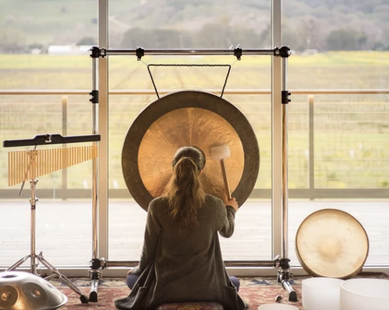 A person sits indoors facing a large gong, hitting it with a mallet. Other musical instruments are nearby. Large windows show a scenic field and hills outside.